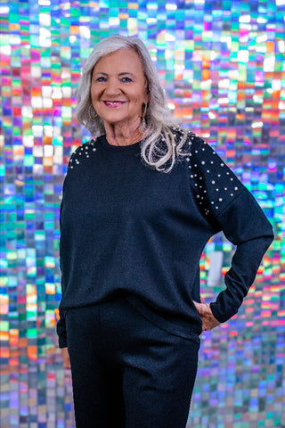 A smiling older woman with shoulder-length gray hair stands against a colorful mosaic tile wall, wearing a dark navy long-sleeve top with white polka dots on the shoulders and dark pants.