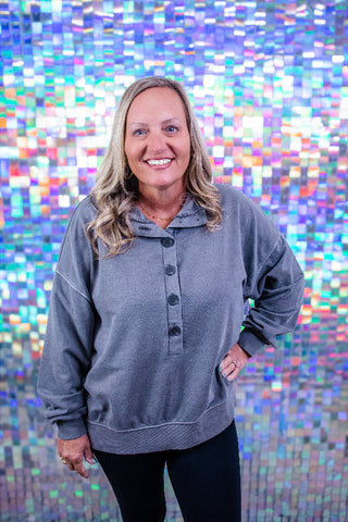 A smiling blonde woman wearing a gray long-sleeve henley sweatshirt and black pants, standing in front of a colorful holographic mosaic tile wall.