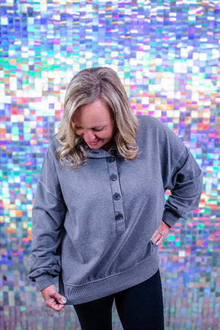 A woman wearing a gray long-sleeve henley sweatshirt with black buttons, standing in front of a colorful, reflective pixelated background.