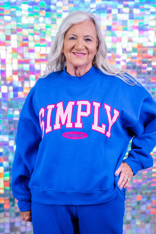 An older woman with gray hair is wearing a bright blue sweatshirt with 'SIMPLY' in white letters outlined in pink, and a small pink oval logo below. She is standing in front of a colorful, reflective mosaic tile wall.