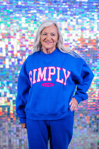 An older woman with gray hair stands smiling in front of a colorful, reflective mosaic tile wall. She is wearing a bright blue sweatshirt with 'SIMPLY' printed in pink and white arched letters across the chest, paired with matching blue sweatpants.