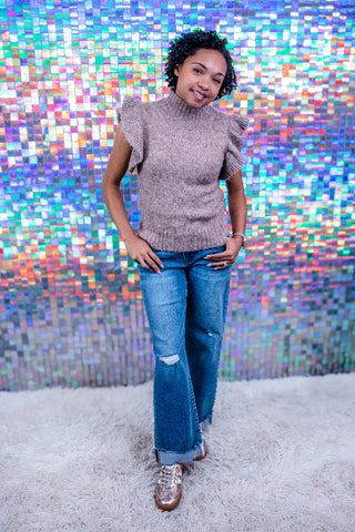A woman modeling a taupe ribbed knit mock-neck sweater with ruffled cap sleeves, paired with blue jeans and brown lace-up shoes, standing on a white shag rug in front of a colorful holographic mirror backdrop.
