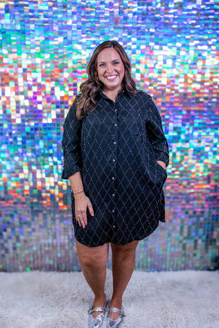 A woman modeling a black button-down shirt dress with a white geometric diamond pattern, standing in front of a colorful, reflective mosaic tile wall.