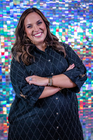 A smiling woman with long brown hair stands with arms crossed, wearing a black button-front shirt dress with a white dotted geometric pattern. She is posed in front of a colorful, reflective mosaic tile wall.