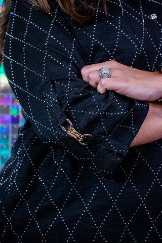 Close-up of a black quilted garment with a white dotted diamond pattern, featuring a gold-tone horsebit clasp on the cuff and a hand adjusting the sleeve.