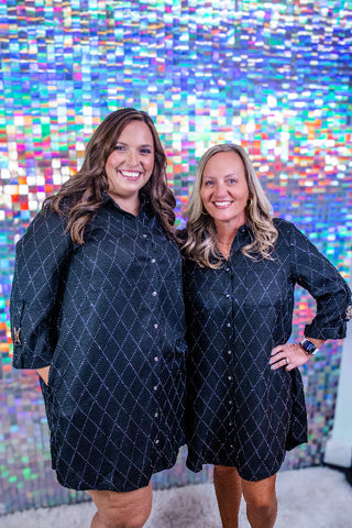 Two women standing side by side wearing matching black button-front shirt dresses with a subtle white geometric pattern, posed in front of a colorful, reflective mosaic wall.