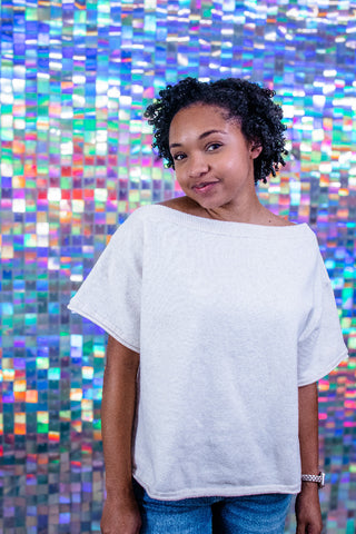A woman with curly dark hair wearing a light gray heather off-the-shoulder t-shirt and blue jeans, standing in front of a colorful holographic mosaic tile wall.