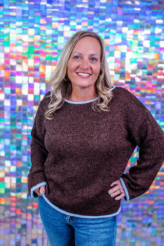 A smiling blonde woman wearing a dark brown textured knit sweater with light blue trim and blue jeans, standing in front of a colorful mosaic tile wall.