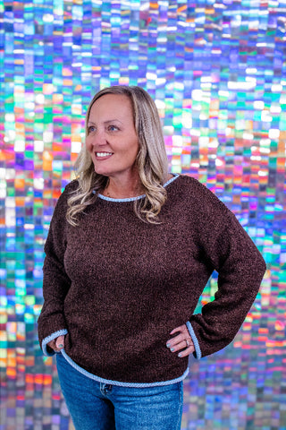 A smiling blonde woman wearing a dark brown textured knit sweater with light blue trim, paired with blue jeans, standing in front of a colorful, pixelated mosaic background.