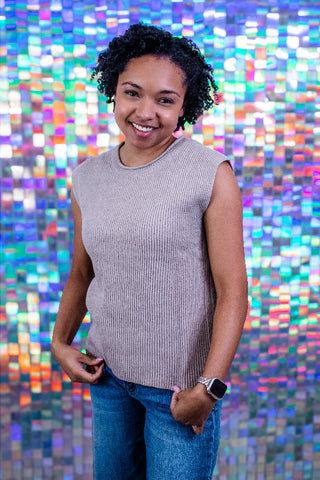 A smiling woman with curly black hair stands in front of a colorful mosaic tile wall, wearing a sleeveless ribbed knit top in a light taupe-beige color and blue jeans.