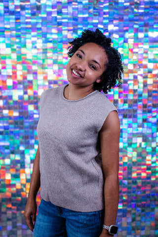 A woman with curly black hair smiling and looking to the side, wearing a sleeveless ribbed knit top in a light grayish-brown color and blue jeans, standing in front of a colorful holographic mosaic tile wall.