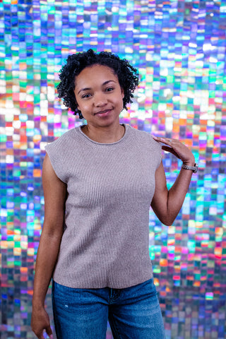 A woman with short curly dark hair wearing a sleeveless ribbed knit top in a light taupe-gray color and blue jeans, posing in front of a vibrant, multicolored pixelated mosaic background.