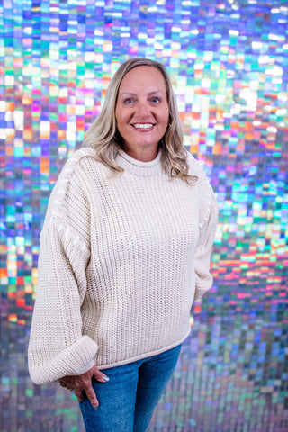 A smiling blonde woman wearing a chunky cream-colored knit sweater and blue jeans, standing in front of a colorful, reflective mosaic wall.