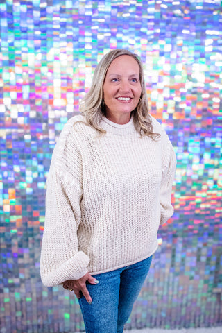 A smiling blonde woman wearing a chunky cream knit sweater and blue jeans, standing in front of a colorful holographic tile wall.