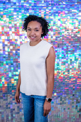 A woman with curly black hair stands in front of a colorful, reflective mosaic tile wall, wearing a white ribbed sleeveless top and blue jeans.