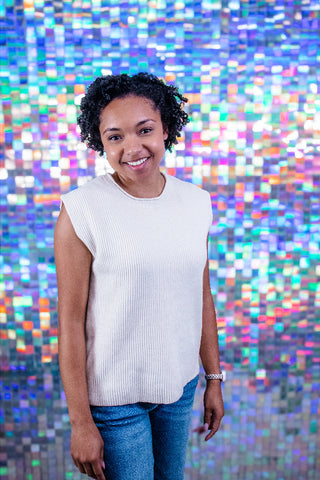 A smiling woman with curly dark hair stands in front of a colorful holographic mosaic wall, wearing a sleeveless ribbed white top and blue jeans.