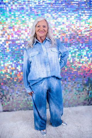A woman with gray hair wearing a light blue denim button-up shirt and matching wide-leg jeans, standing in front of a colorful, reflective mosaic wall.