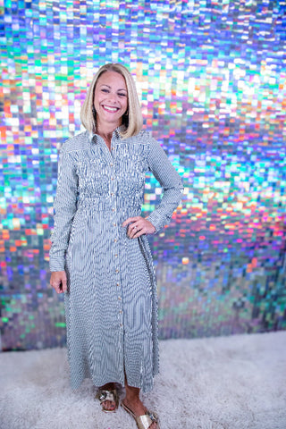 A smiling blonde woman stands in front of a colorful holographic mirror backdrop wearing a long-sleeve, button-front midi dress with a subtle vertical stripe pattern in light blue and white. She is wearing brown and gold flat sandals on a white shag rug.