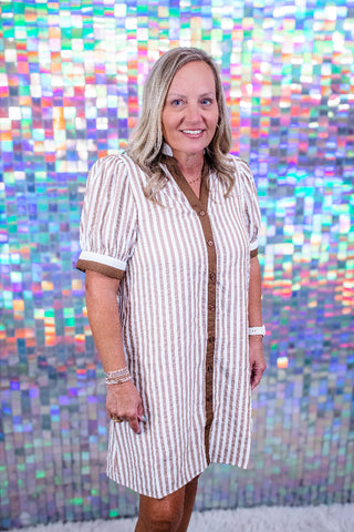 A woman with blonde hair stands in front of a colorful, iridescent mosaic tile wall, wearing a short-sleeve button-up shirt dress with vertical brown and cream stripes.