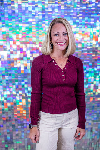 A smiling blonde woman wearing a burgundy ribbed long-sleeve henley top and light beige pants, standing in front of a colorful holographic mosaic wall.