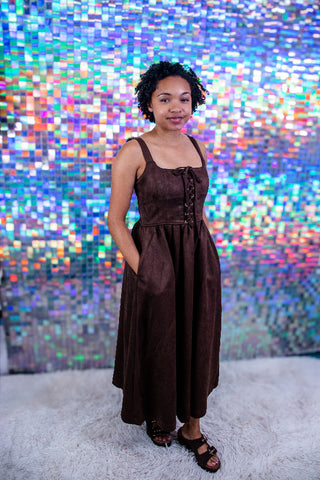 A woman wearing a dark brown sleeveless midi dress with a lace-up front and side pockets, standing on a white shag rug in front of a colorful, reflective mosaic tile wall.