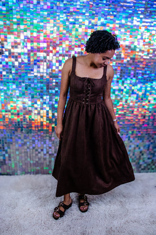 A woman wearing a dark brown sleeveless midi dress with a corset-style bodice and front lace-up detail, standing on a white shag rug in front of a colorful mirrored wall.