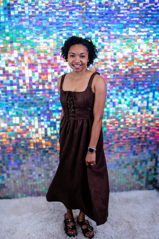 A woman wearing a dark brown sleeveless midi dress with a lace-up front and corset-style bodice, standing in front of a colorful, pixelated mosaic wall.