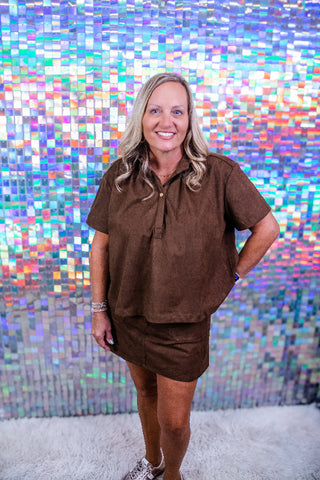 A woman with blonde hair wearing a brown short-sleeve collared shirt and matching brown skirt, standing in front of a colorful holographic tile wall.