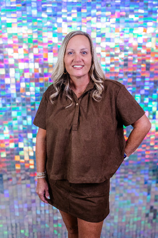 A woman with blonde hair wearing a brown short-sleeve button-up shirt and matching brown skirt, standing in front of a colorful, reflective mosaic tile wall.