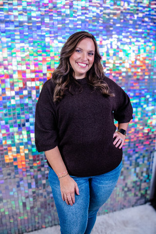 A woman with long brown hair smiling in front of a colorful, reflective mosaic tile wall. She is wearing a dark brown knit mock-neck sweater and blue jeans.