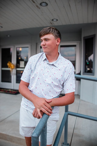 A young man wearing a white short-sleeve button-up shirt with a tiny red and blue pattern, paired with white shorts, leaning on a blue-gray metal railing in front of a building.
