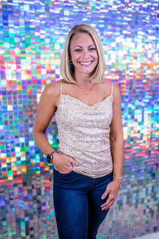 A smiling blonde woman wearing a sparkly champagne sequin camisole top and dark blue jeans, standing in front of a colorful holographic mosaic tile wall.