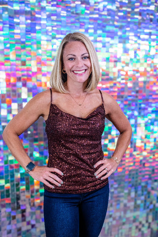 A smiling blonde woman stands with hands on hips wearing a sparkly dark brown sequined camisole top and blue jeans, posed in front of a colorful holographic mosaic wall.