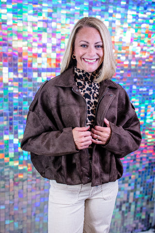 A smiling blonde woman wearing a dark brown suede-like cropped jacket over a leopard-print turtleneck and cream pants, standing in front of a colorful iridescent mosaic tile wall.