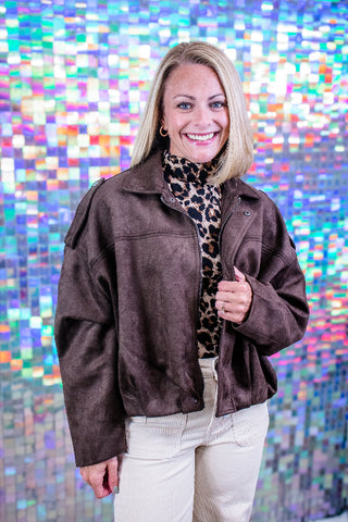 A smiling blonde woman wearing a dark brown suede jacket over a leopard print turtleneck and cream pants, standing in front of a colorful holographic mirror background.