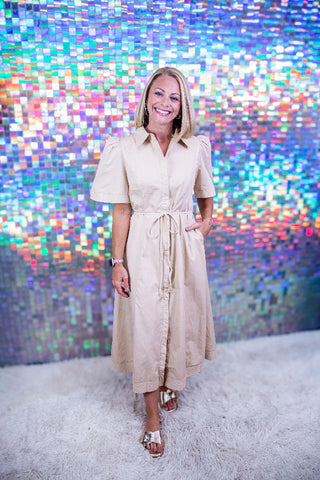 A woman modeling a beige short-sleeve midi shirt dress with a tie waist and front buttons, standing on a white shag rug in front of a colorful, pixelated mirror backdrop.