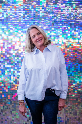 A woman with blonde hair wearing a light blue button-down shirt and dark jeans, standing in front of a colorful, reflective mosaic tile wall.