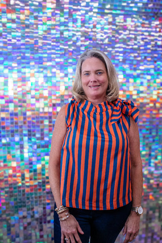 A woman with blonde hair stands in front of a colorful mosaic tile wall, wearing a sleeveless top with vertical orange and navy blue stripes and dark pants.