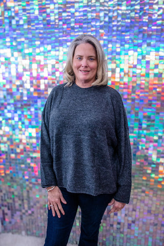 A woman with blonde hair wearing a dark gray long-sleeve knit sweater and dark jeans, standing in front of a colorful, reflective mosaic tile wall.