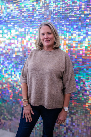 A woman with blonde hair wearing a beige heathered knit top and dark jeans, standing in front of a colorful, reflective mosaic wall.