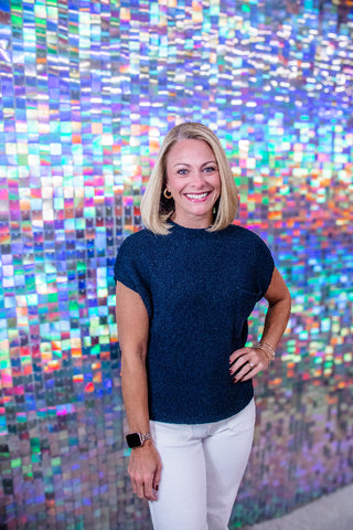 A smiling blonde woman stands in front of a colorful mirrored mosaic wall, wearing a dark navy short-sleeve knit top and white pants.