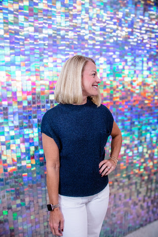 A woman with blonde hair wearing a dark navy blue short-sleeve knit top and white pants, standing in front of a colorful, reflective mosaic tile wall.