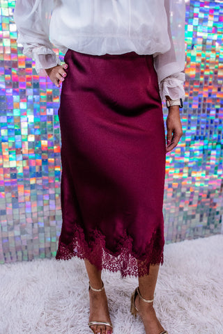 A person wearing a satin-like burgundy midi skirt with lace trim at the hem, paired with a white blouse and metallic heeled sandals, standing in front of a colorful mirrored wall and white rug.