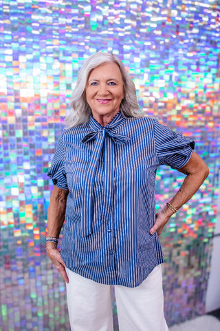 A woman with gray hair wearing a blue and white vertically striped short-sleeve blouse with a bow tie collar and white pants, standing in front of a colorful, reflective mosaic tile wall.