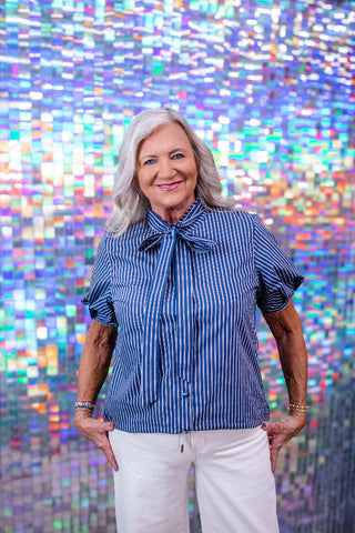 An older woman with gray hair stands smiling in front of a colorful, reflective mosaic wall. She is wearing a blue and white vertically striped short-sleeve blouse with a bow tie collar and white pants.