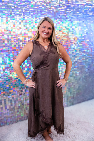 A woman wearing a sleeveless brown maxi dress with lace trim and a tie waist, standing in front of a colorful holographic mirror background.