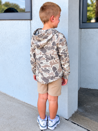 A young boy stands with his back to the camera wearing a beige and brown camouflage hoodie, khaki shorts, and white sneakers with blue accents, standing outdoors against a light gray stucco wall.