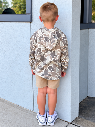 A young boy stands with his back to the camera wearing a beige and brown camouflage hooded sweatshirt, khaki shorts, and white sneakers, posing against a light blue-gray stucco building wall.