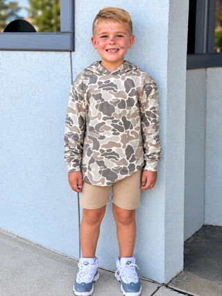 A young boy stands against a light blue-gray stucco wall wearing a long-sleeve camouflage hoodie in beige, brown, and gray tones, paired with tan shorts and white and blue sneakers.