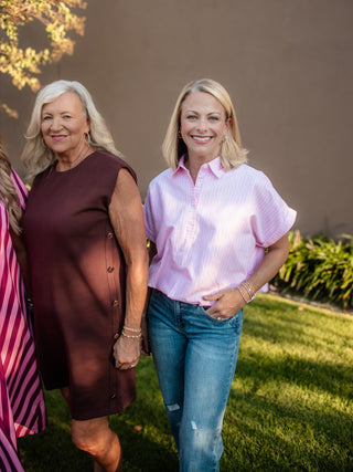 Two women standing outdoors on a grassy lawn in front of a brown wall. The woman on the left wears a sleeveless dark brown dress, and the woman on the right wears a light pink short-sleeve button-up shirt with blue jeans.